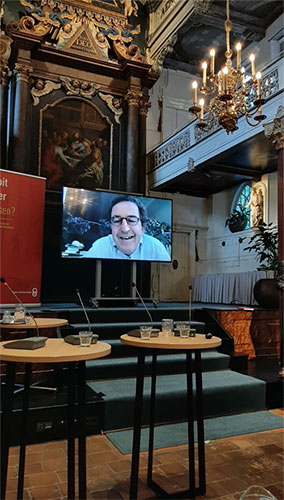 Foto van de zaal met Diederik Gommers op een groot beeldscherm.